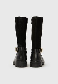 Par črnih škornjev do kolen z usnjenim spodnjim delom in zgornjim delom iz semiša, s podrobnostjo zlatega zaponka in zadrgo na zadnji strani.