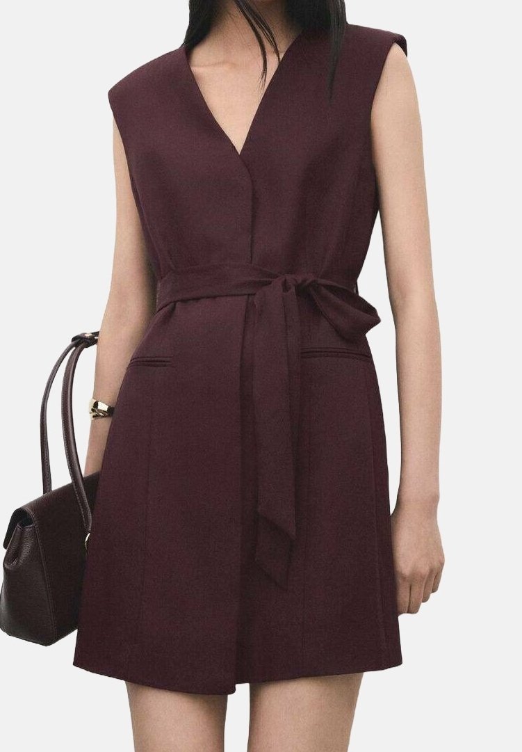 Robe bordeaux sans manches avec col en V et ceinture à la taille, portée par une personne tenant un sac à main bordeaux assorti sur le bras gauche.
