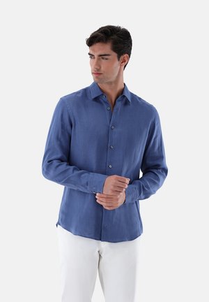 Blaues Langarmshirt aus leichtem Stoff, mit Kragen zum Knöpfen und geradem Saum, kombiniert mit weißen Hosen.