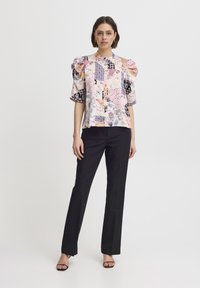 Blusa floreale con maniche a sbuffo corte, colletto alto e vari motivi colorati, abbinata a pantaloni neri su misura e tacchi.