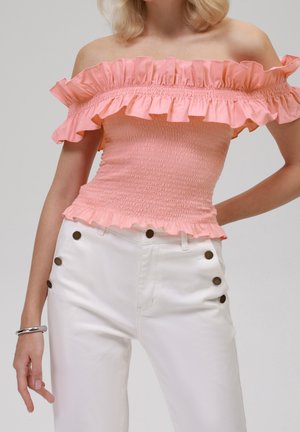 Kobieta w różowej bluzce na ramiączkach z falbanami oraz w białych spodniach z wysokim stanem, z trzema ciemnymi guzikami po każdej stronie, srebrna bransoletka na lewym nadgarstku.