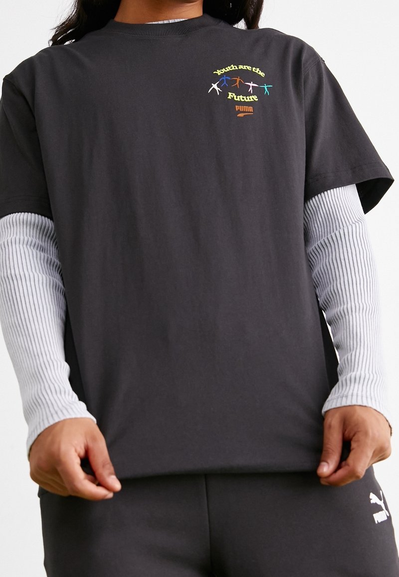 Osoba oblečená v černém oblečení Puma s dlouhými bílými rukávy pod tričkem s nápisem "Mladí jsou budoucnost" nad barevnými siluetami lidí.