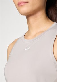 Harmaa istuva hihaton urheilu toppi, jossa on pyöreä kaula-aukko ja hillitty Nike-merkki valkoisena. Materiaalina on sileä ja joustava kangas.