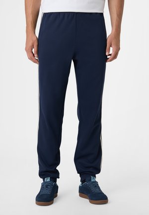 Man draagt marineblauwe trainingsbroek met witte strepen aan de zijkant en marineblauwe sneakers met patroonveters op een grijze achtergrond.