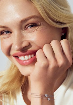 Sølvarmbånd med to hjerteformede charms, en med glitrende detaljer, båret på en hånd der hviler mod en lys farvet top.