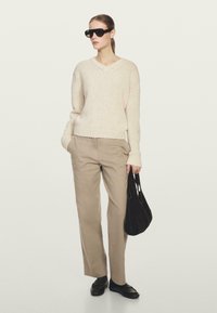 Beige gebreide v-hals truie, losse crèmekleurige broek, zwarte oversized zonnebril en een donkere schoudertas. Platte zwarte schoenen maken de look compleet.