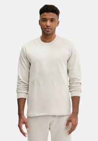 Felpa beige chiaro con collo rotondo, maniche lunghe e un tessuto leggermente testurizzato. Presenta un logo Adidas tenue sul petto.