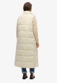 Gilet lungo puffer beige chiaro con design a cuciture orizzontali, colletto alto e fessure laterali. Indossato sopra un maglione di lana crema e jeans blu.