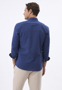 Camisa de manga larga azul marino con puños abotonados, textura suave, corte entallado y un ligero curvado en el dobladillo, combinada con pantalones beige.