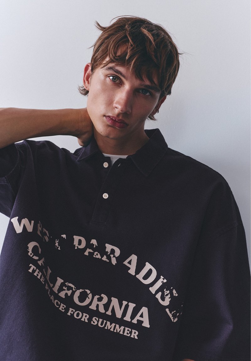 Jeune homme aux cheveux bruns décoiffés, portant un polo sombre avec le texte "California", main reposée sur le cou, sur fond clair uni.