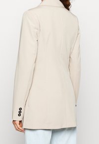 Kvinna iklädd en beige skräddarsydd blazer med svarta knappar på ärmarna, i kombination med ljusblå denimjeans, sedd bakifrån.