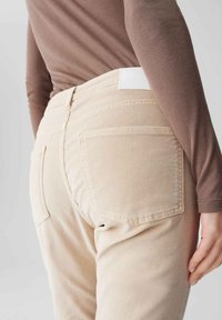 Lichtbeige, middellange broek gemaakt van zachte stof, met achterzakken en een wit label op de tailleband. Soepele textuur en optimaal pasvorm.