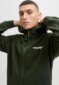 Haut de sweat à capuche zippé vert foncé en tissu texturé, avec des accents côtelés aux épaules et un logo sur la poitrine gauche. Comprend un col haut.