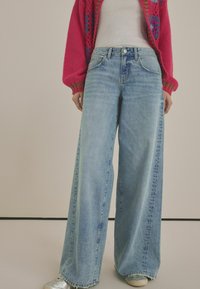 Široké modré džíny z denimu s světlým umytím, mají vysoký pas, viditelné švy a přední zapínání na knoflík.