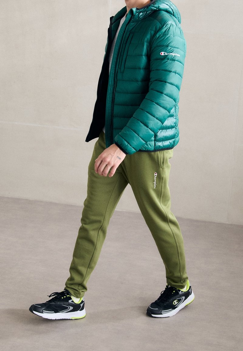 Personne marchant vêtue d'une doudoune bleu sarcelle, d'un pantalon de survêtement vert olive et de baskets noires avec des accents blancs et néon, sur un sol uni.