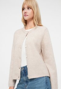 Světle béžový pletený kardigan s kulatým výstřihem, pěti knoflíky vpředu a žebrovanými manžetami, doplněný bílým topem a džíny.