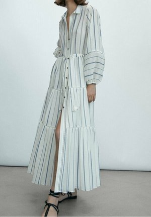 Donna che indossa un lungo vestito bianco con righe verticali blu, chiusura con bottoni sul davanti, maniche a sbuffo e sandali neri con cinturini.