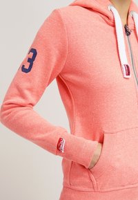 Felpa con zip color corallo, con lacci bianchi, tasca a marsupio, numero 3 navy sulla manica e toppa rossa del brand sul polsino.