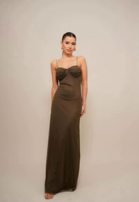 Olivno zelena maxi obleka z tankimi naramnicami, s nabranim trupom in tekočo teksturo. Model stoji ob nevtralni ozadju.