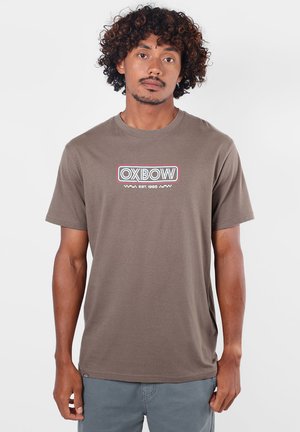 Homme aux cheveux bouclés portant un t-shirt marron OXBOW et un pantalon gris, debout sur un fond blanc uni.