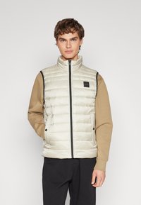 Helle beige Steppweste mit hohem Kragen, Reißverschluss auf der Vorderseite, zwei seitlichen Taschen und dunkelbraunen Akzenten. Solides, gesteppte Textur mit Logo-Patch.