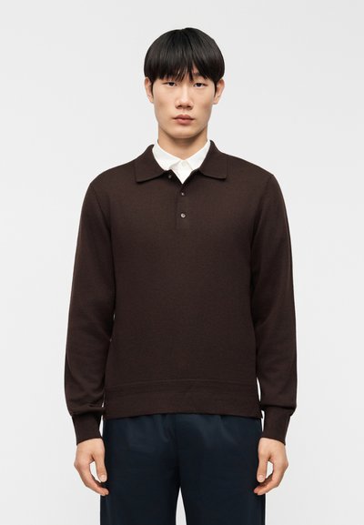 Pull à manches longues marron avec col polo, trois boutons et poignets côtelés. Porté par-dessus une chemise blanche à boutons.