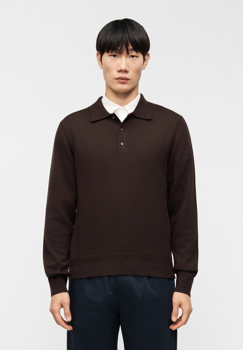 Καφέ μπλούζα πόλο με μακριά μανίκι α, γιακά, τρία κουμπιά και ριμπ μανσέτες. Φοριέται πάνω από λευκό πουκάμισο με κουμπιά.