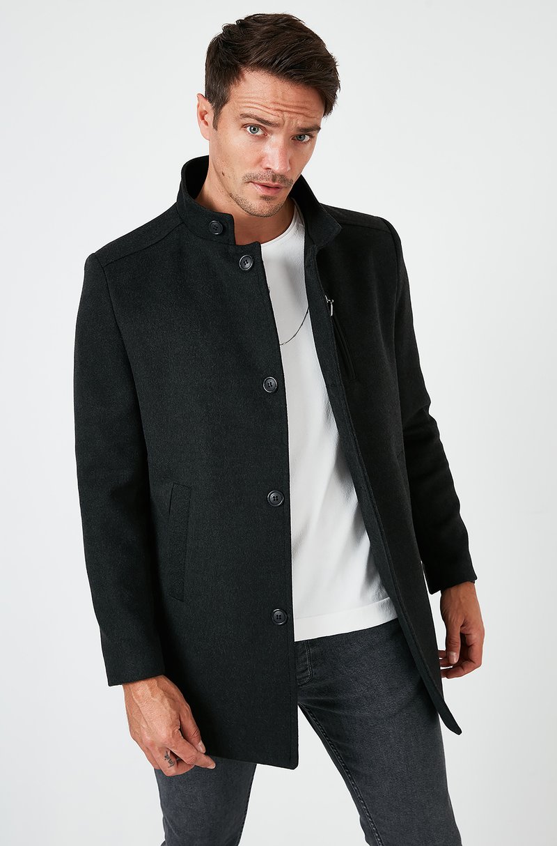 Abrigo de mezcla de lana gris oscuro con un cuello alto, botones frontales y bolsillos laterales. Lleva puesto sobre una camisa blanca y jeans negros. Textura suave.