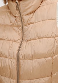 Béžová prošívaná vesta s vysokým límcem, horizontálním prošíváním a středovým zipem s kovovým jezdcem. Měkká, hladká textura.