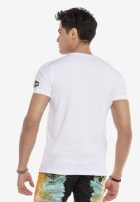 Das weiße, figurbetonte T-Shirt verfügt über einen Rundhalsausschnitt und kurze Ärmel. Es hat ein kleines gesticktes Logo auf der linken Schulter. Der Stoff ist weich und glatt.
