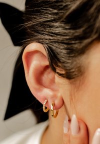 Pendientes de aro de oro, uno con gemas claras y el otro pulido, expuestos en la oreja de un modelo. Acabado suave y brillante; detalle en primer plano.