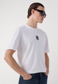 T-shirt en coton blanc à manches courtes, avec un petit logo graphique noir sur la poitrine. Assorti à un jean en denim foncé.