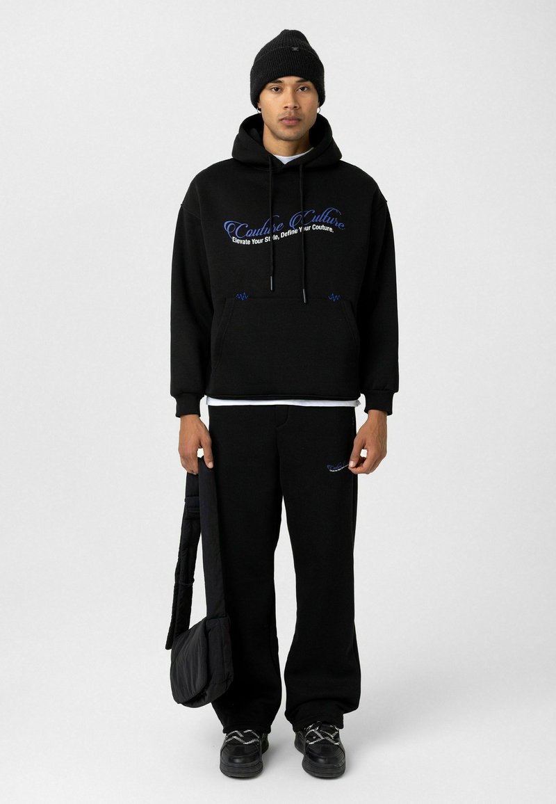 Zwarte hoodie met blauwe borduring, met een voorzak en trekkoorden. Matte zwarte wide-leg trainingsbroek, gecombineerd met zwarte sneakers en een tas.