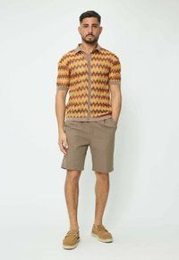 Chemise à manches courtes avec un motif en zigzag rouge, jaune et marron. Associée à un short beige et des chaussures à enfiler marron.