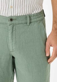 Pantaloni in lino verde chiaro con design a vita piatta, cintura elastica e tasca laterale. Il tessuto ha una finitura morbida e testurizzata.