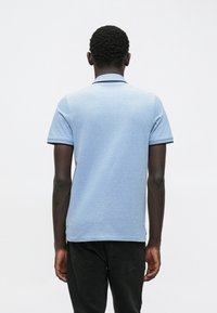 Homme aux cheveux courts et bouclés portant un polo bleu clair à manches courtes et un pantalon noir, debout de dos sur un fond uni.