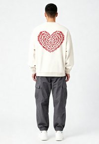 Crèmekleurige sweatshrit met een grote rode hartgrafiek van tekst op de achterkant, gecombineerd met grijze cargobroek. De stof lijkt zacht en heeft een ontspannen pasvorm.