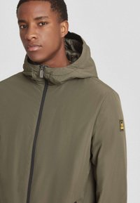 Chaqueta con capucha de color verde oliva, hecha de material ligero, con cierre de cremallera y una franja negra de acento, con la marca en la manga.