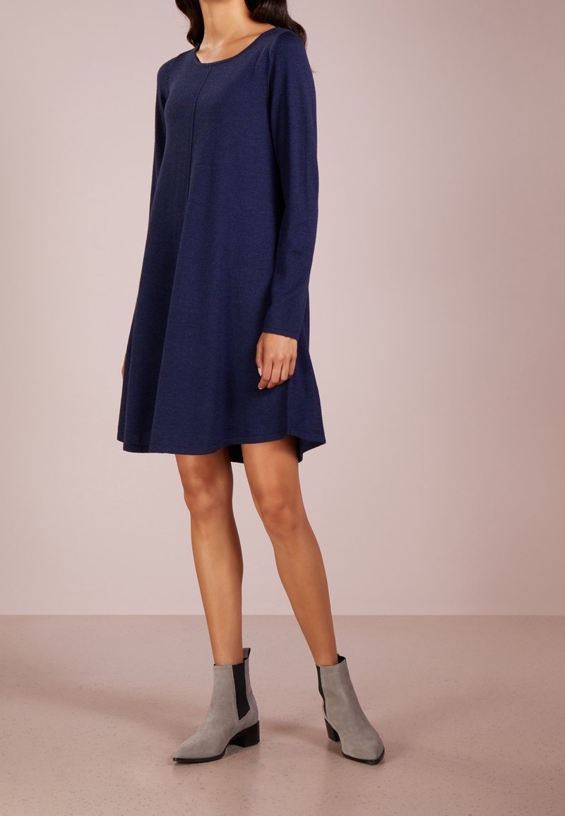 Femme portant une robe ample à manches longues bleu marine et des bottines gris clair, debout devant un fond uni.