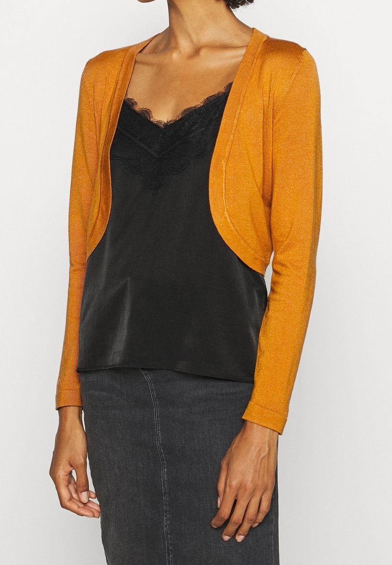 Oranger schimmernder Cardigan mit offener Vorderseite, langen Ärmeln und einem glatten schwarzen Spitzen-Top darunter, kombiniert mit einem dunklen Jeansrock.