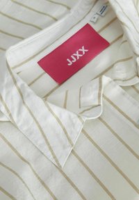 Camisa a rayas en tejido color crema y beige, con cuello, tapeta de botones y una etiqueta rosa con la inscripción "JJXX". Material texturizado y ligero.
