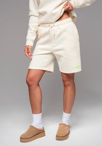 Shorts en coton couleur crème avec une taille élastique et un cordon de serrage, arborant un logo vert fluo sur la jambe inférieure gauche.
