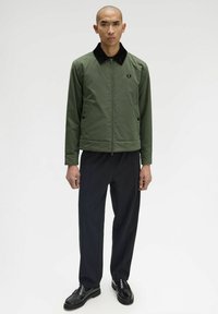 Giacca verde con zip e colletto nero, dotata di tasche laterali e un logo. Indossata con pantaloni neri e calzature.