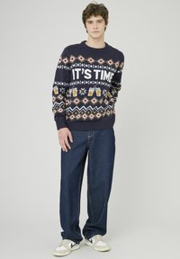 Pull en tricot bleu marine avec des motifs festifs blancs et dorés, arborant le texte « IL EST TEMPS », associé à un jean en denim noir décontracté et des baskets blanches.
