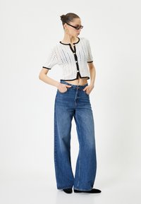 Weißes gestricktes Cropped-Top mit schwarzem Saum und Knöpfen, kombiniert mit weit geschnittenen blauen Jeans. Das Model steht mit den Händen in den Taschen und trägt eine Sonnenbrille.