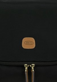 Zaino in nylon nero con cerniere dorate, rifiniture in pelle marrone e un’etichetta con logo rettangolare. Tessuto liscio e design elegante.