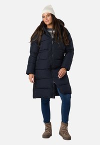 Lang marineblå dunjakke med horisontale syninger, frontlynlås og hætte. Kombineret med denimjeans og brune termiske støvler. Strikket cremefarvet beanie-hat.