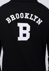 Black sweatshirt featuring "BROOKLYN" arched in white letters and a large white letter "B" on the back. Soft cotton texture.