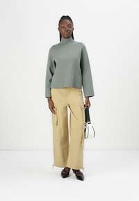Lichtgroene coltrui, beige wijde cargo-broek met zakken en zwarte punten-schoenen. Minimalistische zwart-witte handtas.
