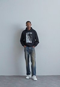 Sudadera negra con capucha y gráfico en el frente, combinada con jeans de mezclilla azul claro y zapatillas blancas, sobre un fondo neutro.